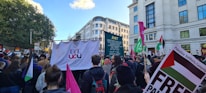 A large crowd gathers on a street for a protest or demonstration. They are holding various signs and banners, including a prominent one displaying 'UCL UCU' and others with phrases related to a campaign. Many participants hold flags and placards, and the scene is set against an urban backdrop with tall buildings and trees in the distance.