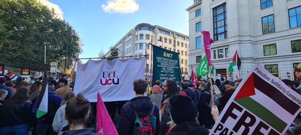 A large crowd gathers on a street for a protest or demonstration. They are holding various signs and banners, including a prominent one displaying 'UCL UCU' and others with phrases related to a campaign. Many participants hold flags and placards, and the scene is set against an urban backdrop with tall buildings and trees in the distance.