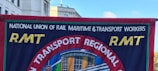 A banner displays the logo and name of the National Union of Rail, Maritime & Transport Workers, known as RMT. The banner's design includes bold yellow letters 'RMT' against a blue background with a central circular emblem showing a building and a blue-red contrast.