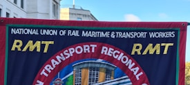 A banner displays the logo and name of the National Union of Rail, Maritime & Transport Workers, known as RMT. The banner's design includes bold yellow letters 'RMT' against a blue background with a central circular emblem showing a building and a blue-red contrast.