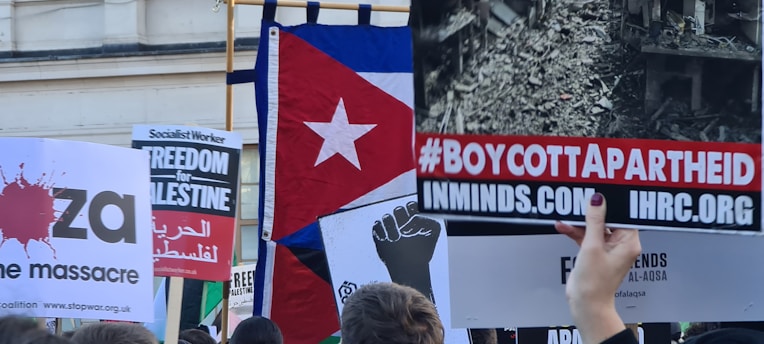 A group of protest signs are held up by a crowd. One sign reads '#BOYCOTTAPARTHEID' with a web address below, while another features a flag with a red star. Additional signs mention 'Freedom for Palestine' and 'Stop the massacre', with slogans in Arabic and English. The image includes a monochrome photograph of destruction and various activist symbols.