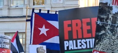 A collection of protest signs and flags, including a Cuban flag and one with 'FREE PALESTINE' text. There is a sign indicating 'Freedom for Palestine' and an image of destruction. The setting appears to be outside a building, with a partially visible street sign in the background.