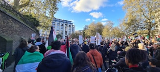 A large group of people gathers outdoors, participating in a protest or rally. The crowd holds various signs and flags, with messages emphasizing peace and freedom. Trees with green leaves and a blue sky with clouds form the backdrop, alongside a large building. The atmosphere is lively and engaged.