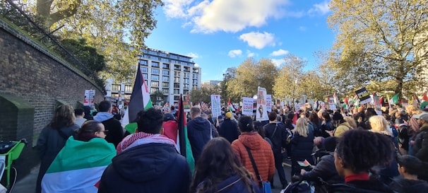 A large group of people gathers outdoors, participating in a protest or rally. The crowd holds various signs and flags, with messages emphasizing peace and freedom. Trees with green leaves and a blue sky with clouds form the backdrop, alongside a large building. The atmosphere is lively and engaged.