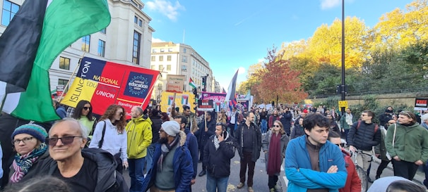 A large group of people are gathered for a protest or demonstration on a city street. They are holding various banners and flags with messages suggesting advocacy for different causes. The scene is set in an urban environment with buildings in the background and trees with autumn-colored leaves. The crowd consists of individuals dressed in casual, cool-weather attire.
