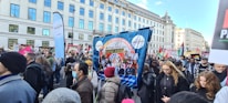 A large group of people is participating in a street protest. They hold various banners and placards with messages related to education and social issues. The crowd is diverse, with some people wearing masks. The protest takes place in an urban setting with tall buildings in the background.