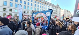A large group of people is participating in a street protest. They hold various banners and placards with messages related to education and social issues. The crowd is diverse, with some people wearing masks. The protest takes place in an urban setting with tall buildings in the background.