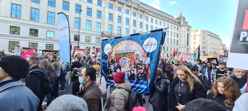 A large group of people is participating in a street protest. They hold various banners and placards with messages related to education and social issues. The crowd is diverse, with some people wearing masks. The protest takes place in an urban setting with tall buildings in the background.