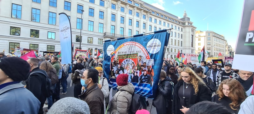 A large group of people is participating in a street protest. They hold various banners and placards with messages related to education and social issues. The crowd is diverse, with some people wearing masks. The protest takes place in an urban setting with tall buildings in the background.