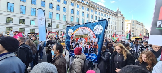 A large group of people is participating in a street protest. They hold various banners and placards with messages related to education and social issues. The crowd is diverse, with some people wearing masks. The protest takes place in an urban setting with tall buildings in the background.