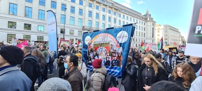 A large group of people is participating in a street protest. They hold various banners and placards with messages related to education and social issues. The crowd is diverse, with some people wearing masks. The protest takes place in an urban setting with tall buildings in the background.