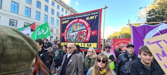 A group of people are participating in a public demonstration. They are carrying various banners and signs, including one prominently displaying 'RMT London Transport Regional Council' and the word 'Socialism'. The demonstrators are gathered in an urban environment with buildings and trees visible in the background.