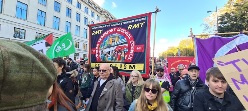 A group of people are participating in a public demonstration. They are carrying various banners and signs, including one prominently displaying 'RMT London Transport Regional Council' and the word 'Socialism'. The demonstrators are gathered in an urban environment with buildings and trees visible in the background.