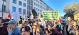 A large gathering of people is seen protesting outdoors in an urban setting. Many of the participants are holding signs and flags. One prominent banner reads 'International Jewish Anti-Zionist Network.' The crowd includes individuals of various ages and appearances, and they seem engaged and focused. The background features a large building and trees, indicating a city environment. The atmosphere appears active and purposeful.