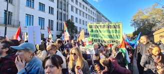 A large gathering of people is seen protesting outdoors in an urban setting. Many of the participants are holding signs and flags. One prominent banner reads 'International Jewish Anti-Zionist Network.' The crowd includes individuals of various ages and appearances, and they seem engaged and focused. The background features a large building and trees, indicating a city environment. The atmosphere appears active and purposeful.