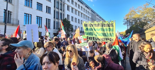 A large gathering of people is seen protesting outdoors in an urban setting. Many of the participants are holding signs and flags. One prominent banner reads 'International Jewish Anti-Zionist Network.' The crowd includes individuals of various ages and appearances, and they seem engaged and focused. The background features a large building and trees, indicating a city environment. The atmosphere appears active and purposeful.