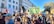 A large gathering of people is seen protesting outdoors in an urban setting. Many of the participants are holding signs and flags. One prominent banner reads 'International Jewish Anti-Zionist Network.' The crowd includes individuals of various ages and appearances, and they seem engaged and focused. The background features a large building and trees, indicating a city environment. The atmosphere appears active and purposeful.