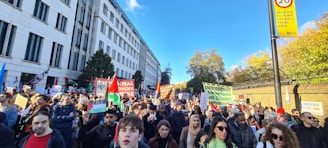 A large group of people is gathered outdoors, holding signs and banners, participating in a protest or demonstration. The scene is lively with a mix of individuals showing solidarity and expressing their opinions. The background includes a tall building and trees.