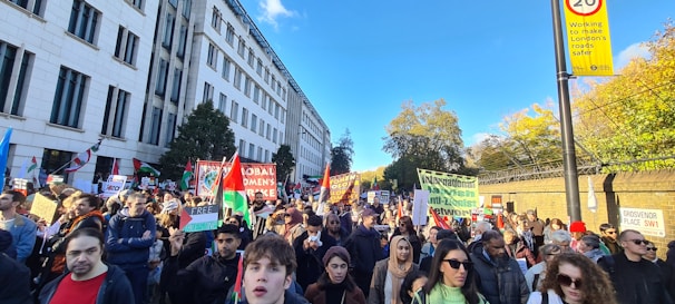 A large group of people is gathered outdoors, holding signs and banners, participating in a protest or demonstration. The scene is lively with a mix of individuals showing solidarity and expressing their opinions. The background includes a tall building and trees.