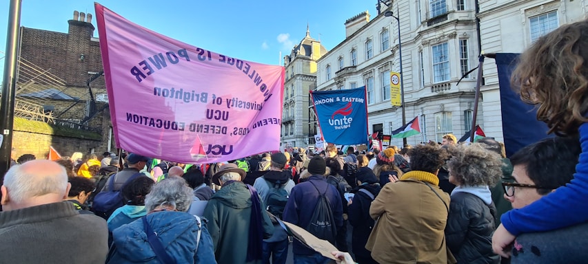 A large group of people is gathered outdoors in what appears to be a protest or rally. They are holding banners and placards with slogans related to education and labor rights. The crowd includes people of different ages and the background consists of historic buildings under a clear blue sky.