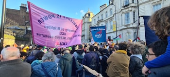 A large group of people is gathered outdoors in what appears to be a protest or rally. They are holding banners and placards with slogans related to education and labor rights. The crowd includes people of different ages and the background consists of historic buildings under a clear blue sky.