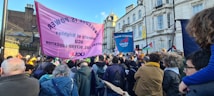 A large group of people is gathered outdoors in what appears to be a protest or rally. They are holding banners and placards with slogans related to education and labor rights. The crowd includes people of different ages and the background consists of historic buildings under a clear blue sky.
