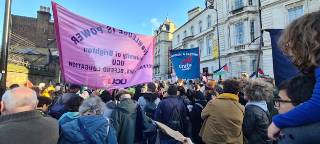 A large group of people is gathered outdoors in what appears to be a protest or rally. They are holding banners and placards with slogans related to education and labor rights. The crowd includes people of different ages and the background consists of historic buildings under a clear blue sky.