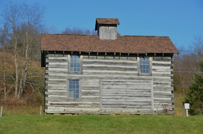 A rustic, two-story wooden log cabin with a brown shingle roof, set against a backdrop of bare trees and open sky. The cabin has several windows with clear glass, and a weathered wagon wheel leans against its side. The structure sits on a grassy lawn, suggesting a rural setting.
