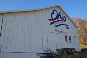 A white wooden barn with a large painting commemorating Ohio's bicentennial event, featuring the word 'Ohio' in blue with a red star and a red and blue ribbon design. It also displays the years '1803' and '2003'. The ground around the barn is grassy with trees in the background displaying autumn colors.