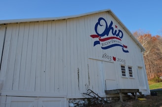 A white wooden barn with a large painting commemorating Ohio's bicentennial event, featuring the word 'Ohio' in blue with a red star and a red and blue ribbon design. It also displays the years '1803' and '2003'. The ground around the barn is grassy with trees in the background displaying autumn colors.