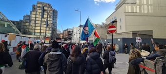 A group of people is participating in a street protest. They are walking along a city street, carrying flags and signs. The flags include one with a peace symbol and another with a national flag design. The crowd is diverse, and some people are wrapped in flags. Buildings surround the area, including a modern glass structure on the left and an older building on the right. A stop sign is visible along the path.