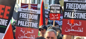 A crowd of people is gathered holding various signs advocating for 'Freedom for Palestine'. The signs are held above the heads of the people, some of whom are partially visible. The text on the signs is bold and prominent, with additional Arabic translations. There are banners in the background, including a red one that reads 'London Eastern'.