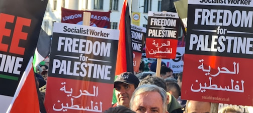 A crowd of people is gathered holding various signs advocating for 'Freedom for Palestine'. The signs are held above the heads of the people, some of whom are partially visible. The text on the signs is bold and prominent, with additional Arabic translations. There are banners in the background, including a red one that reads 'London Eastern'.