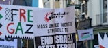 A street scene featuring protest signs with various messages related to political activism. The signs include messages like 'Free Gaza' and 'End Israeli State Terror', indicating a demonstration. The environment appears to be urban with buildings in the background.