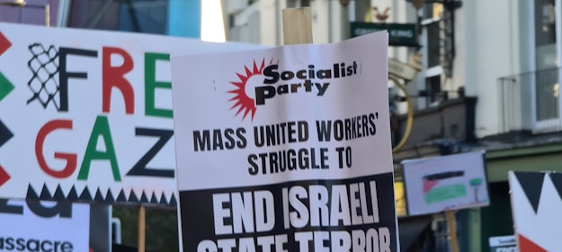 A street scene featuring protest signs with various messages related to political activism. The signs include messages like 'Free Gaza' and 'End Israeli State Terror', indicating a demonstration. The environment appears to be urban with buildings in the background.