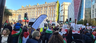 A group of people participates in a street protest, carrying banners and flags. The scene takes place in an urban setting with historic buildings and modern high-rises. Police vehicles are present, and various protest signs focusing on political messages are visible. The crowd is large and densely packed.