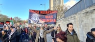 A group of people are participating in a protest, holding signs with strong political messages, including a prominently displayed banner with a photograph and bold text. The scene takes place on a city street lined with trees and tall buildings.