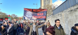 A group of people are participating in a protest, holding signs with strong political messages, including a prominently displayed banner with a photograph and bold text. The scene takes place on a city street lined with trees and tall buildings.