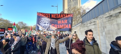 A group of people are participating in a protest, holding signs with strong political messages, including a prominently displayed banner with a photograph and bold text. The scene takes place on a city street lined with trees and tall buildings.
