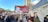 A group of people are participating in a protest, holding signs with strong political messages, including a prominently displayed banner with a photograph and bold text. The scene takes place on a city street lined with trees and tall buildings.