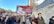 A group of people are participating in a protest, holding signs with strong political messages, including a prominently displayed banner with a photograph and bold text. The scene takes place on a city street lined with trees and tall buildings.