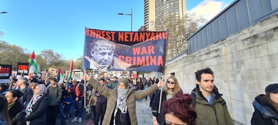 A group of people are participating in a protest, holding signs with strong political messages, including a prominently displayed banner with a photograph and bold text. The scene takes place on a city street lined with trees and tall buildings.