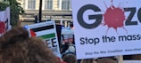 A protest scene with people holding signs advocating for Palestine solidarity and anti-war messages. Several signs feature slogans such as 'Free Palestine' and 'Stop the Massacre.' A police presence is indicated by a visible blue light in the background.