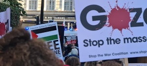 A protest scene with people holding signs advocating for Palestine solidarity and anti-war messages. Several signs feature slogans such as 'Free Palestine' and 'Stop the Massacre.' A police presence is indicated by a visible blue light in the background.
