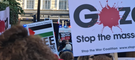 A protest scene with people holding signs advocating for Palestine solidarity and anti-war messages. Several signs feature slogans such as 'Free Palestine' and 'Stop the Massacre.' A police presence is indicated by a visible blue light in the background.