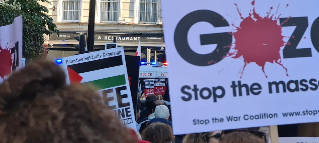 A protest scene with people holding signs advocating for Palestine solidarity and anti-war messages. Several signs feature slogans such as 'Free Palestine' and 'Stop the Massacre.' A police presence is indicated by a visible blue light in the background.