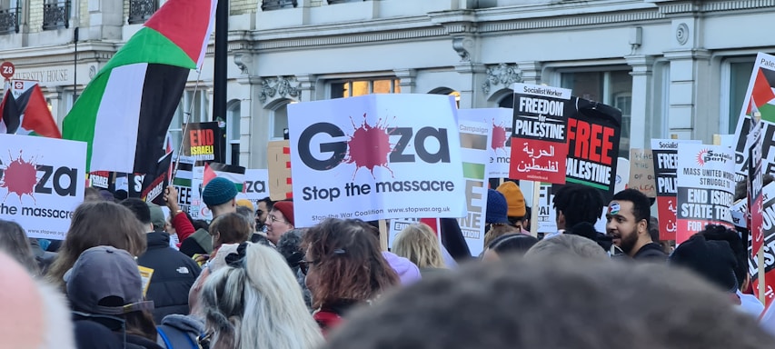 A crowd of people holding various signs and banners supporting Gaza and Palestine. The signs include messages like 'Stop the massacre' and 'Freedom for Palestine.' Some flags are visible, including the Palestinian flag. The setting appears to be a protest or rally in an urban environment.