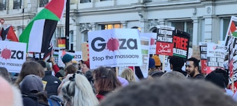 A crowd of people holding various signs and banners supporting Gaza and Palestine. The signs include messages like 'Stop the massacre' and 'Freedom for Palestine.' Some flags are visible, including the Palestinian flag. The setting appears to be a protest or rally in an urban environment.