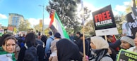 A large group of people are gathered outdoors, holding signs with messages of solidarity. The crowd includes people wearing headscarves and others bundled in winter clothing. There are several signs that read 'Free Palestine', and one person is holding a Palestinian flag. Trees and a building are visible in the background under a blue sky.