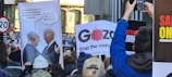 A crowd of protesters holding various signs, including a satirical poster depicting a conversation between two political figures with speech bubbles about bombing and financial support. Other signs include messages and slogans about the situation in Gaza. The scene suggests a public demonstration with participants capturing images.